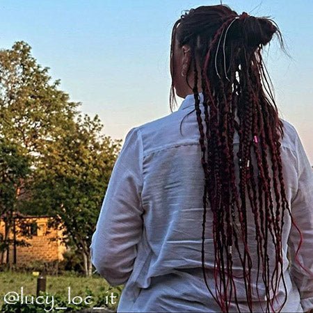 a girl stands with her back against a summer landscape. she has long burgundy synthetic dreadlocks